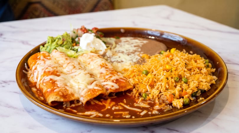 Two cheesy enchiladas with rice, refried beans, guacamole, and sour cream on a brown plate.