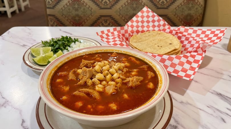 A bowl of menudo soup with hominy, tripe, and a side of limes, onions, and tortillas.