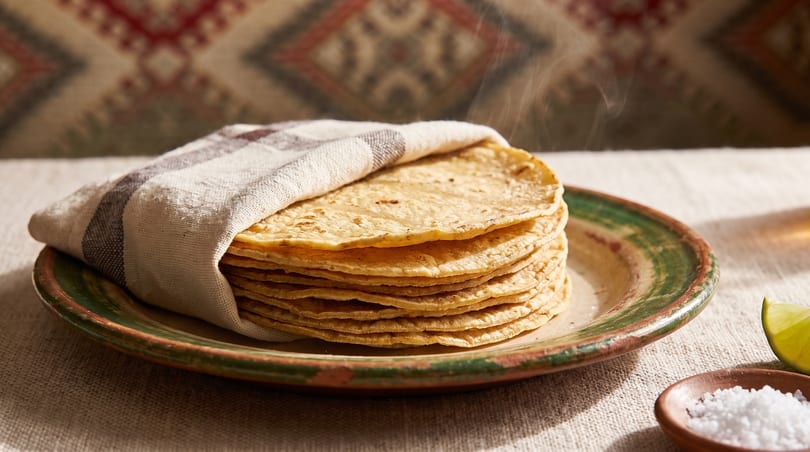 Steaming hot corn tortillas wrapped in a cloth on a rustic plate with salt and lime.