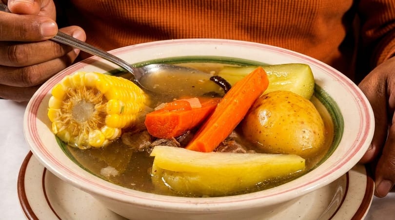 A person eating a bowl of hearty soup with corn on the cob, carrots, potatoes, and other vegetables.