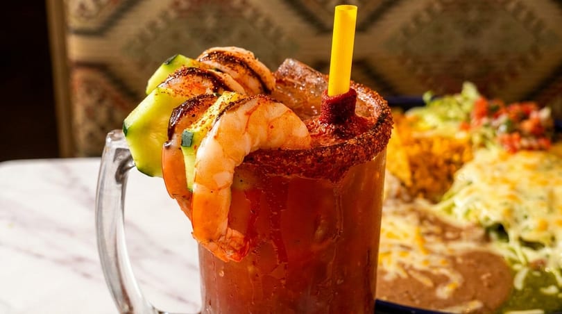 A michelada cocktail with shrimp, cucumber, and a yellow straw, next to a plate of Mexican food.